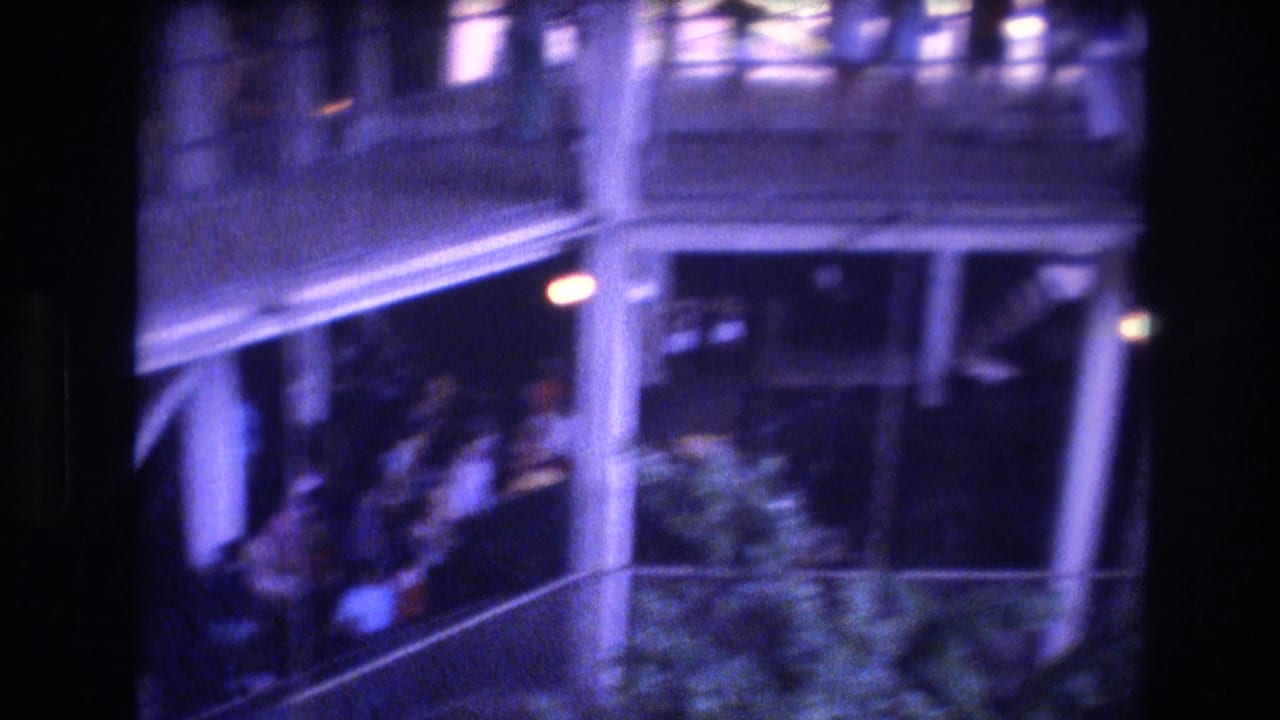 Exterior balcony or walkway with railings, columns, greenery, and blurred people.