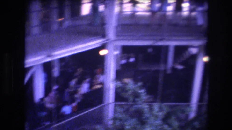 1977 Honolulu 8mm view of a balcony or exterior walkway with railings, vertical supports, tropical greenery, and indistinct people in the background.