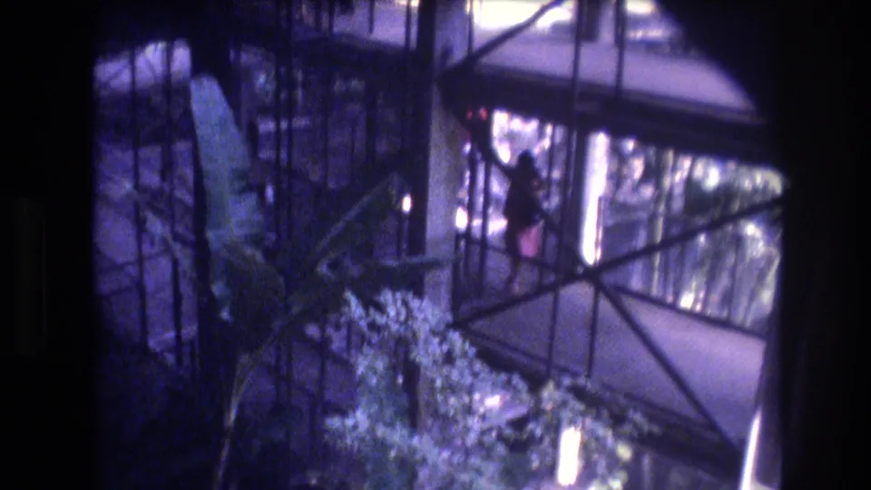 1977 Honolulu home-movie frame showing a glass-enclosed atrium walkway, metal beams, tropical foliage, and a distant person in red on an upper level.