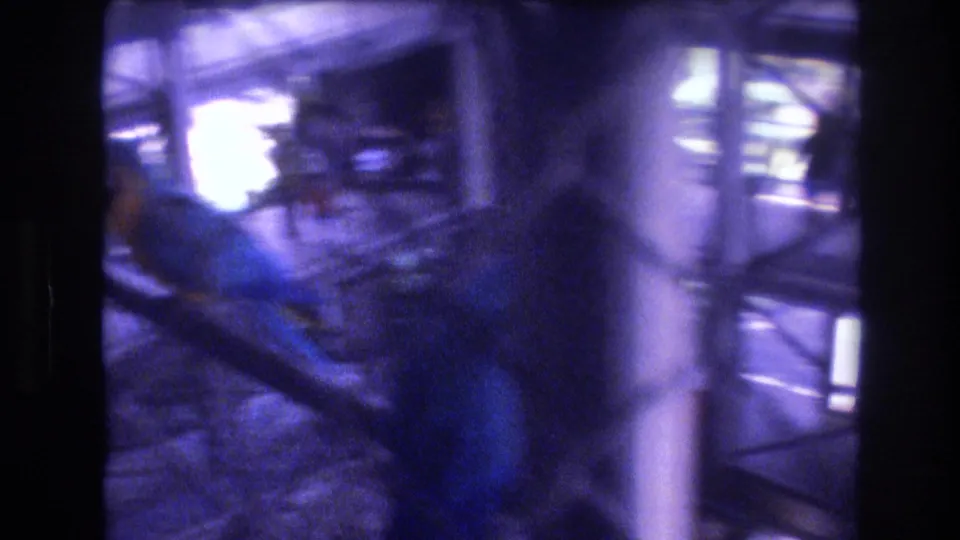 A close handheld view shows the blue-and-yellow macaw inside the zoo enclosure, partly obscured by fencing, posts, and exposure flare.