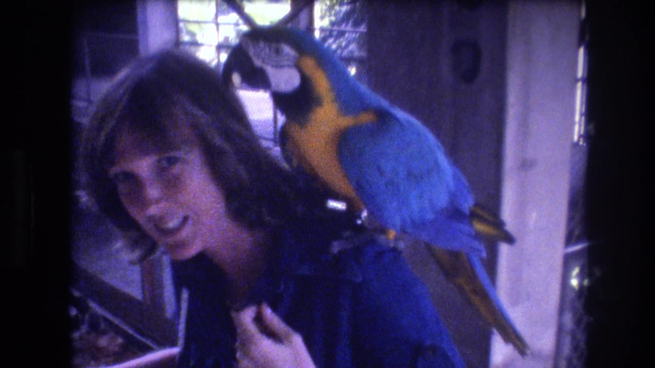 Smiling woman with a blue-and-yellow macaw on her shoulder near a gridded window.