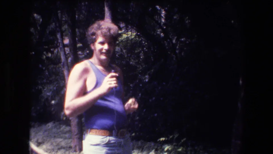 A person wearing a blue tank top and light-colored pants stands outdoors in a wooded area in the 1977 Honolulu home-movie footage.