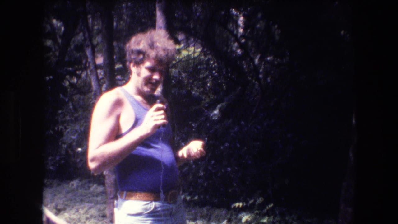 In 1977 Honolulu home-movie footage, a curly-haired man in a purple sleeveless top holds a microphone while standing in a wooded outdoor area.