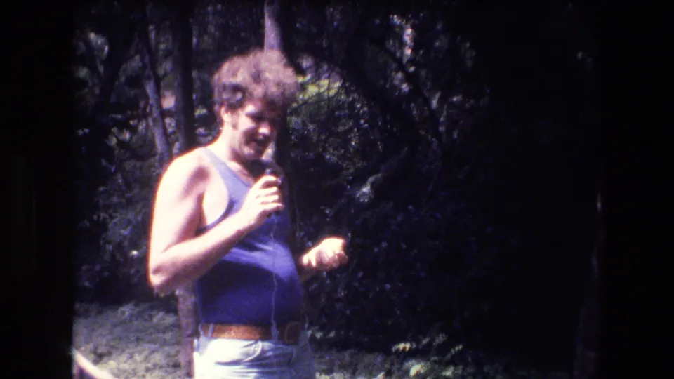 In 1977 Honolulu home-movie footage, a curly-haired man in a purple sleeveless top holds a microphone while standing in a wooded outdoor area.