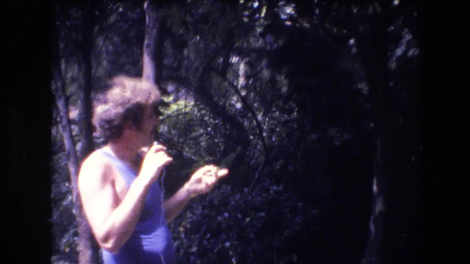 The performer turns in profile, holding a microphone and gesturing with his free hand in a wooded outdoor area identified by metadata as Honolulu in 1977.