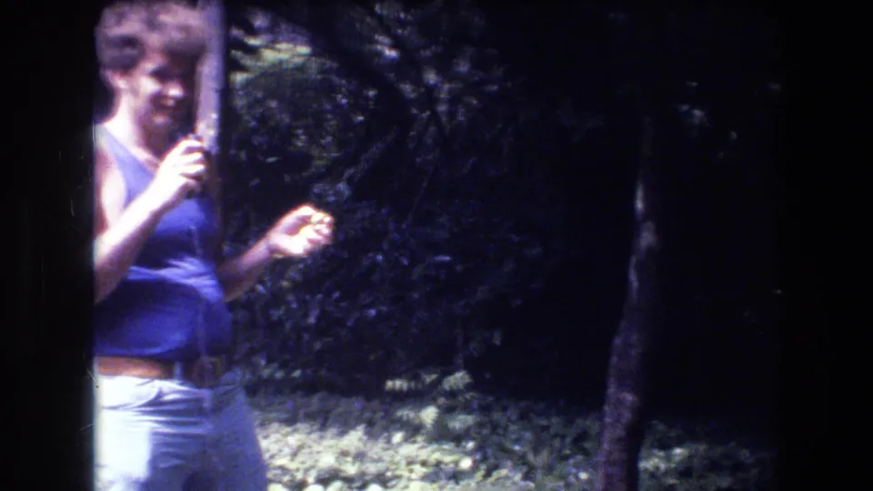 1977 Honolulu-area home-movie frame showing an adult man in a blue tank top and light pants partly cropped at the left edge among dense trees.