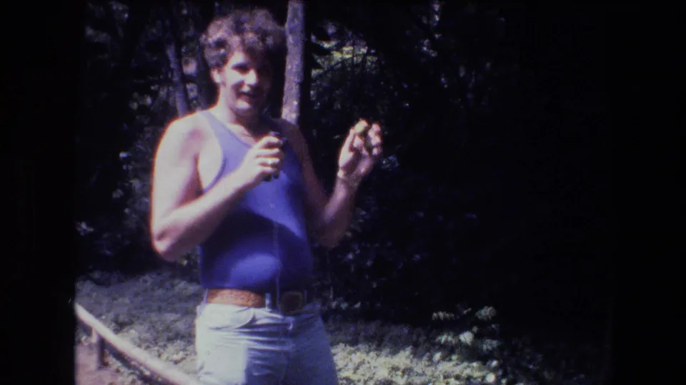 1977 Honolulu-area 8mm frame showing an adult man in a blue tank top and light pants standing near trees while holding a small object in his hands.