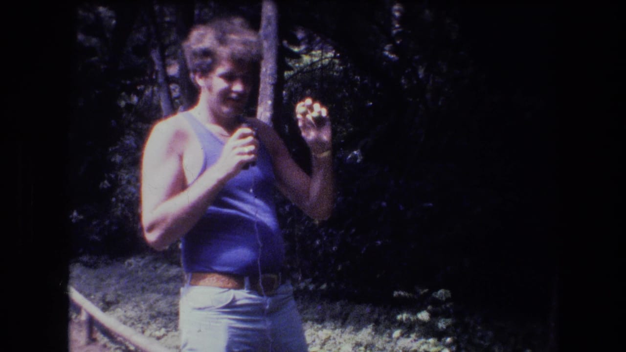 In 1977 Honolulu, Oahu home-movie footage, a curly-haired man in a blue tank top stands in a wooded area and holds a small object near his face.