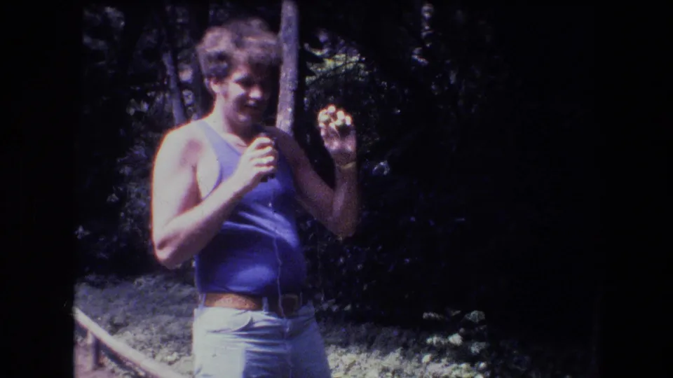 In 1977 Honolulu, Oahu home-movie footage, a curly-haired man in a blue tank top stands in a wooded area and holds a small object near his face.