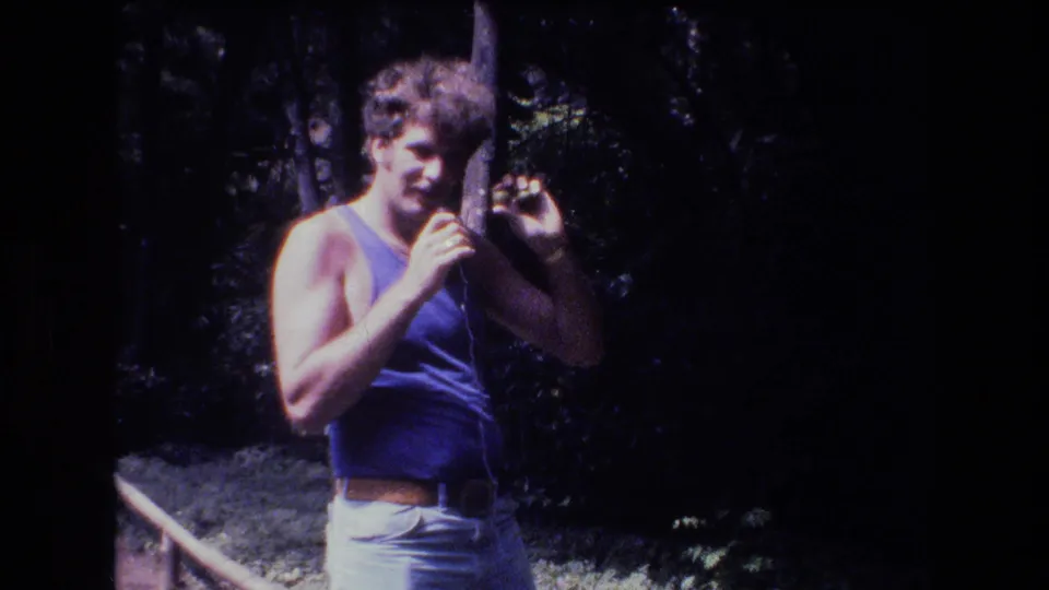 A curly-haired man faces the camera in 1977 home-movie footage from Honolulu, gesturing as if explaining the small object in his raised hand.
