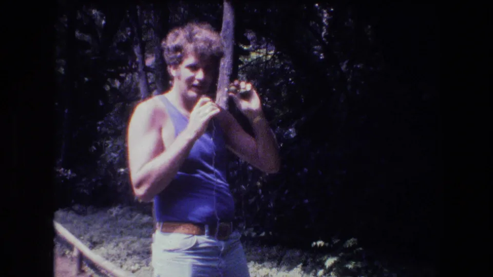 The man remains framed in the trees, holding the small object beside his face as the 8mm image shows heavy contrast and vintage color shift.