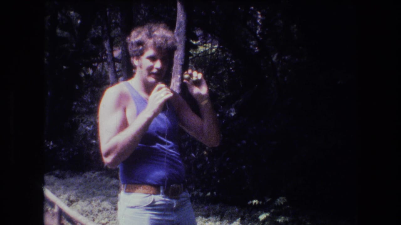 In metadata-supported 1977 Honolulu home-movie footage, a curly-haired man in a blue tank top stands in a shaded wooded area holding a thin wire or stick.
