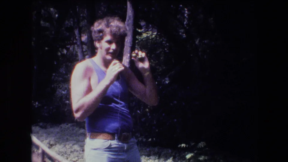 In metadata-supported 1977 Honolulu home-movie footage, a curly-haired man in a blue tank top stands in a shaded wooded area holding a thin wire or stick.