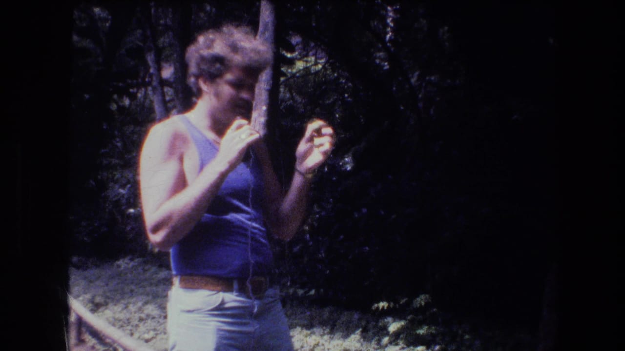 Man in a blue tank top looks downward while holding a thin wire or stick outdoors.