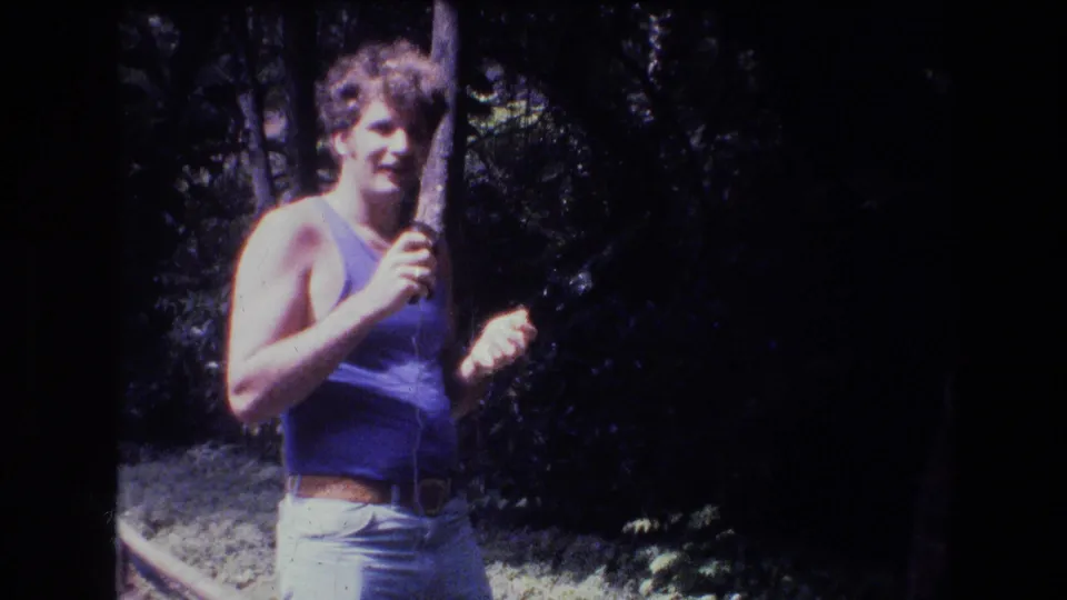 A curly-haired adult man faces the camera in a casual 1977 Honolulu home-movie frame, holding a thin wire or stick with trees and dark foliage behind him.