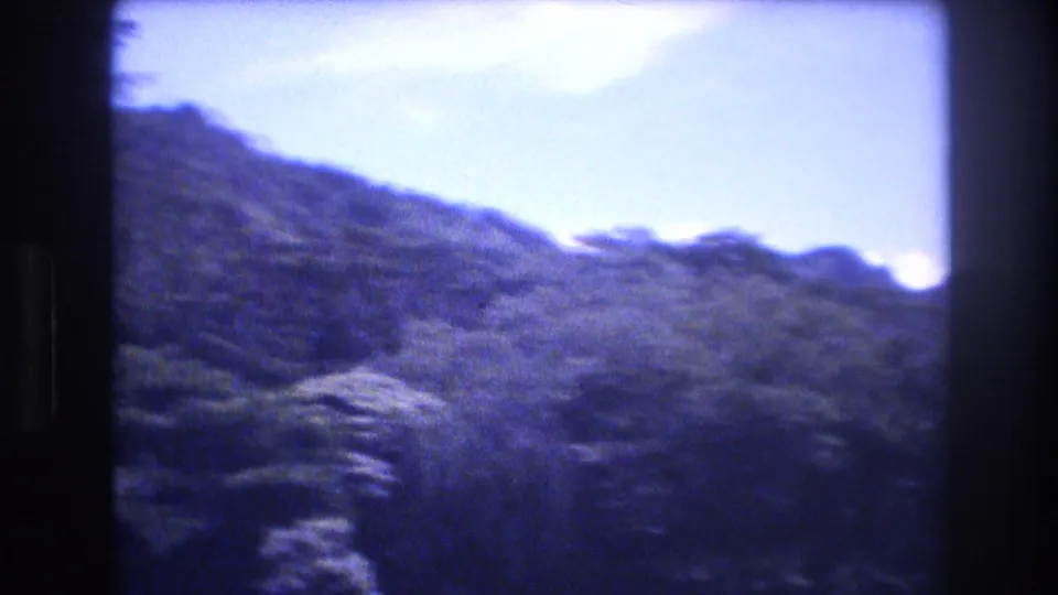 1977 Honolulu and Oahu 8mm frame with a tree-covered mountain slope, pale blue sky, and soft blue-purple film color.