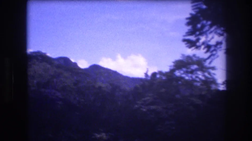 1977 Honolulu and Oahu home-movie frame showing distant mountain silhouettes, treetops, bright blue sky, and small clouds through handheld 8mm framing.