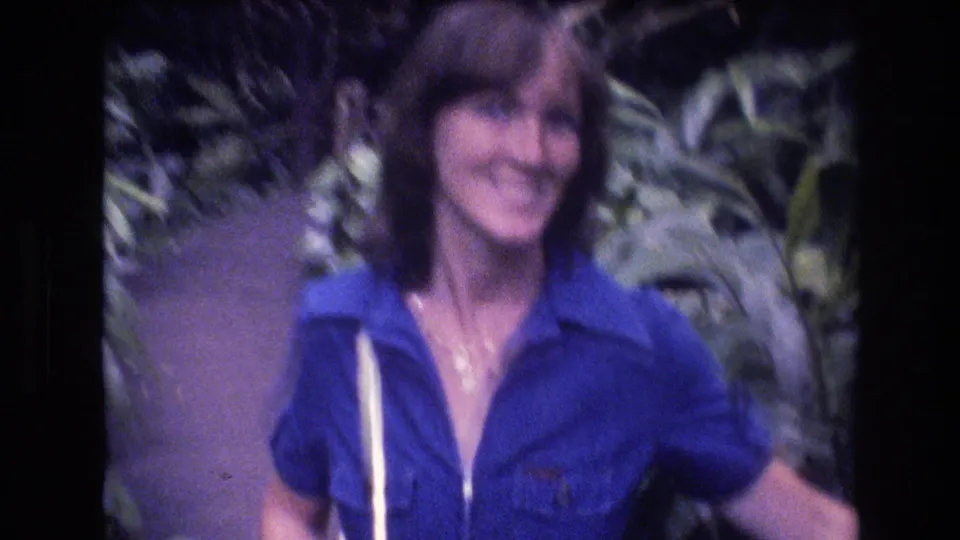 Young woman with brown hair in a bright blue collared shirt smiles toward the camera in 1977 Honolulu, with tropical plants behind her and a white strap over her shoulder.