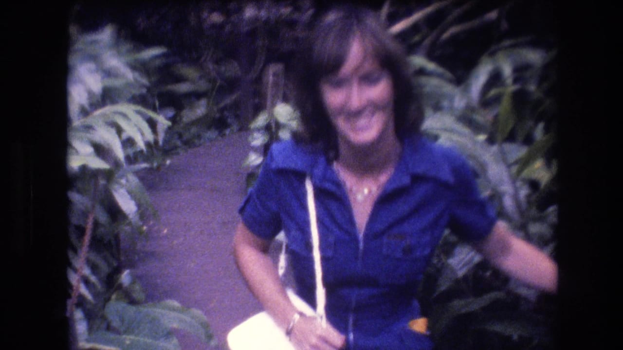Smiling woman in a blue shirt walking on a narrow path through dense tropical plants.