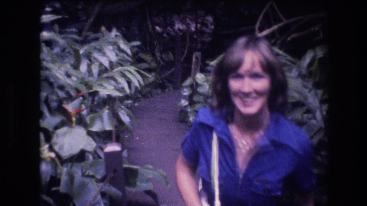 The woman continues along the Oahu rainforest path, smiling toward the handheld camera amid broad leaves and shaded vegetation.