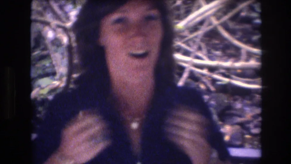 A smiling adult woman in a dark blue top faces the camera with raised hands among branches and foliage in the 1977 Honolulu home-movie footage.