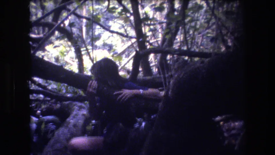 1977 Honolulu Oahu home-movie frame with a seated long-haired figure in a blue top beside fallen wood and thick undergrowth.