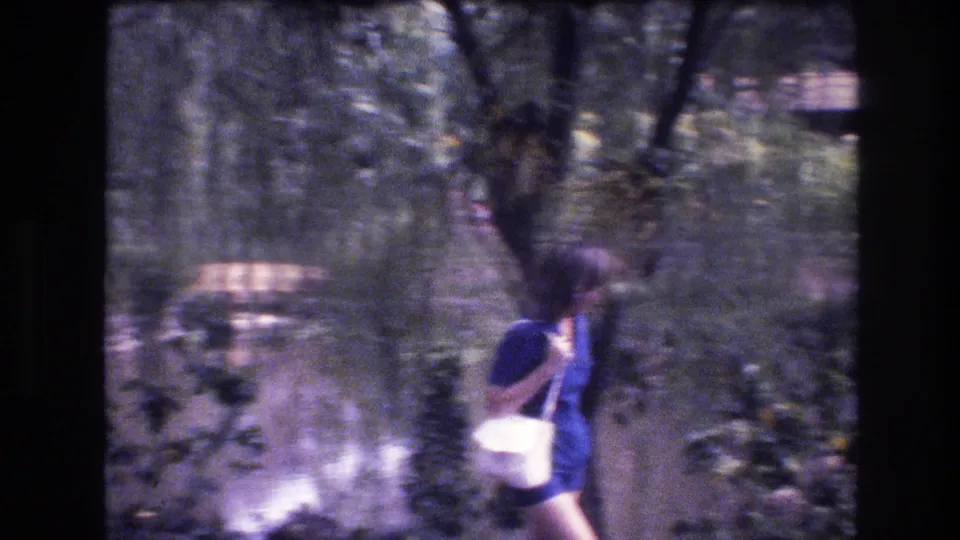 1977 Honolulu, Oahu 8mm frame showing a person in blue clothing carrying a white shoulder bag while walking through a leafy garden area.