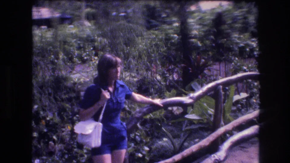 A woman in a blue outfit with a white shoulder pocketbook stands near the rustic bridge in 1977 Honolulu home-movie footage.