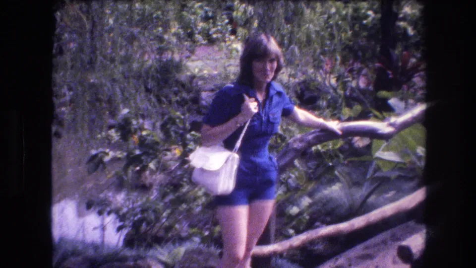 The woman in a blue outfit crosses the rustic wooden bridge with a white shoulder bag in the Honolulu tropical garden scene.