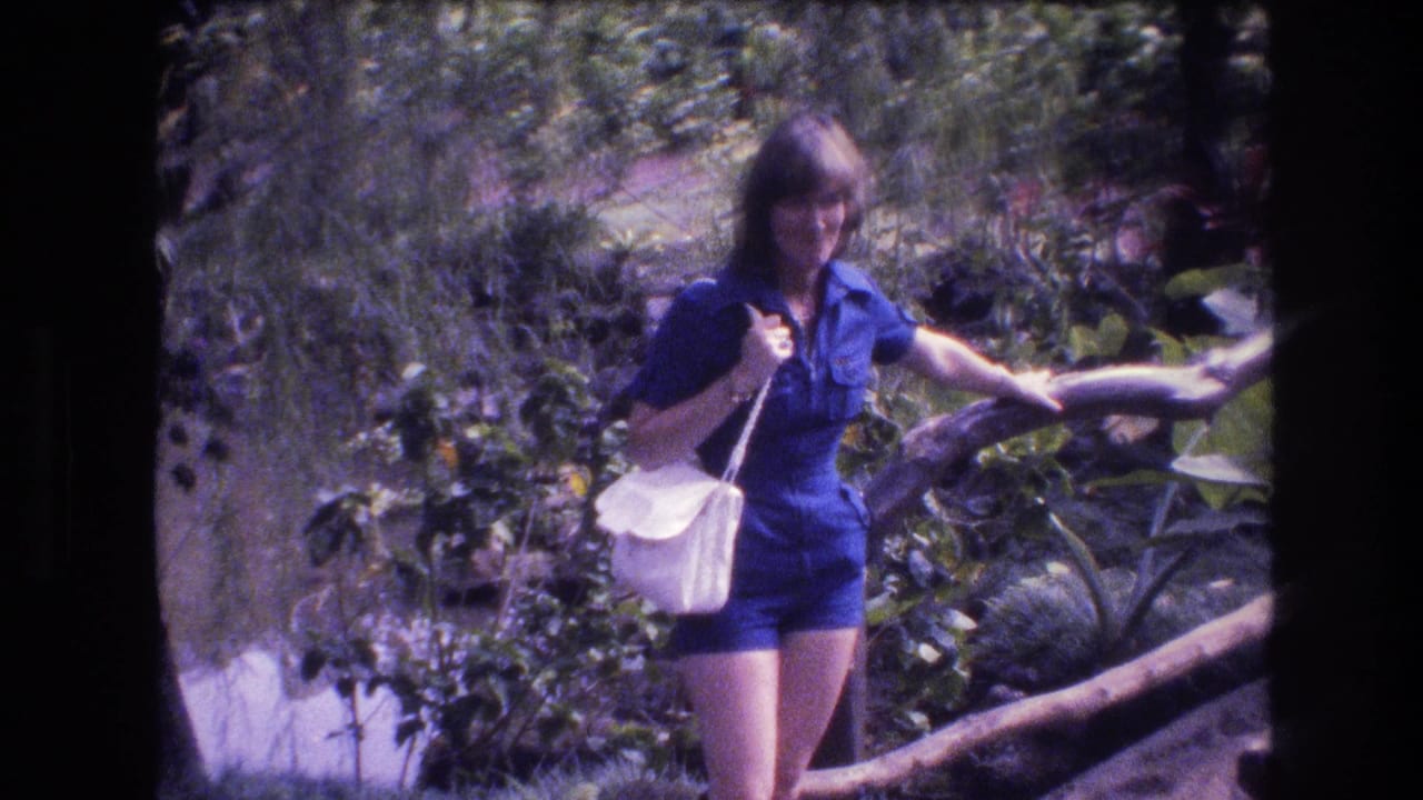 In 1977 Honolulu, Oahu home-movie footage, a woman in a blue romper and white shoulder purse smiles while moving along a wooden railing amid dense tropical greenery.
