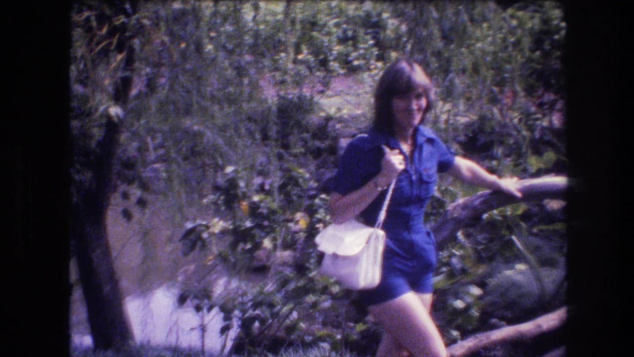 The woman in a blue romper continues through the Honolulu garden setting, partly framed by foliage and a wooden rail, with strong sunlight and soft 8mm color.