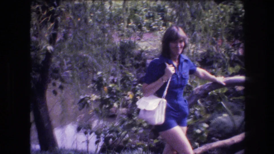 The woman in a blue romper continues through the Honolulu garden setting, partly framed by foliage and a wooden rail, with strong sunlight and soft 8mm color.
