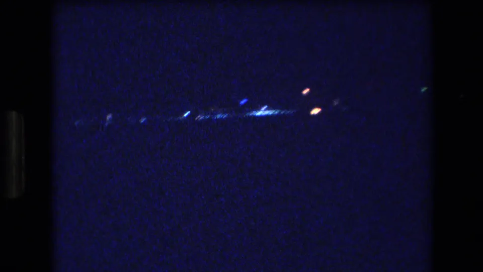 1977 Honolulu or Oahu night frame with a sparse band of glowing lights and heavy 8mm grain in the darkness.