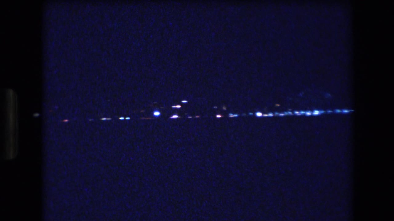 A grainy 1977 Honolulu home-movie frame shows scattered white and blue lights along a dark waterfront horizon beneath a deep blue night sky.