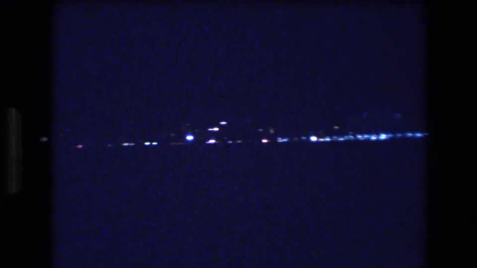 A grainy 1977 Honolulu home-movie frame shows scattered white and blue lights along a dark waterfront horizon beneath a deep blue night sky.
