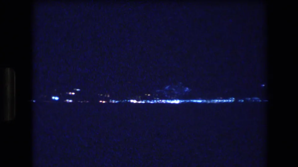 A low-light 8mm frame from Honolulu, Oahu, shows clusters of blue-white lights spread across a distant shoreline over dark water.