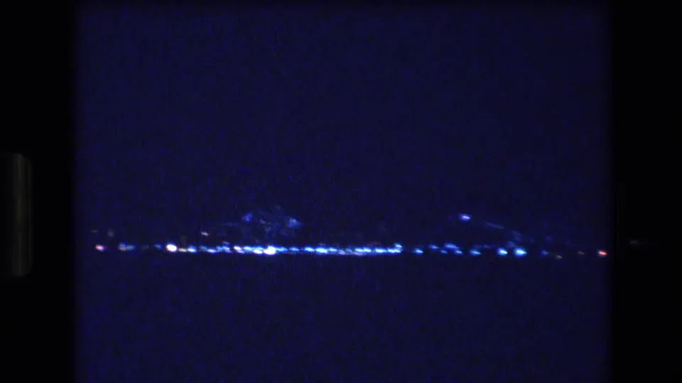 The frame holds on a dim 1977 Honolulu nighttime horizon, with scattered lights and faint reflections barely separating the shoreline from the water.