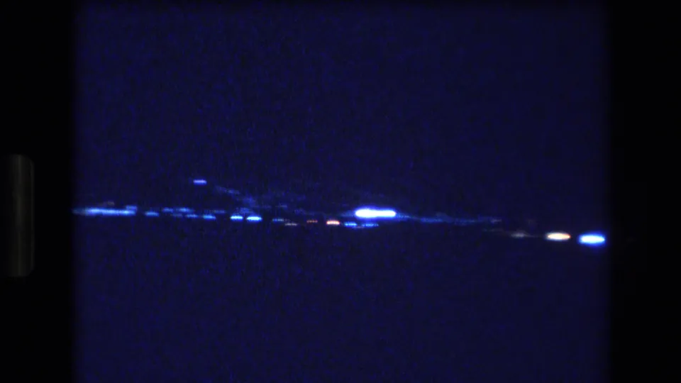A grainy Honolulu night frame shows a brighter cluster of lights toward the right, with a dark expanse of water and a faint shoreline under a blue-black sky.