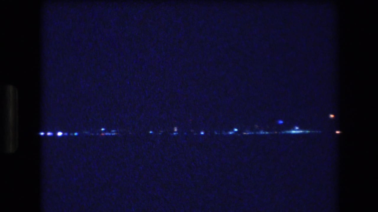 A 1977 Honolulu 8mm night frame shows scattered blue-white lights along a low horizon across dark water, with heavy grain and frame-edge darkness.