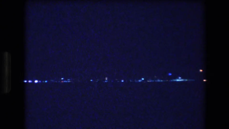 A 1977 Honolulu 8mm night frame shows scattered blue-white lights along a low horizon across dark water, with heavy grain and frame-edge darkness.