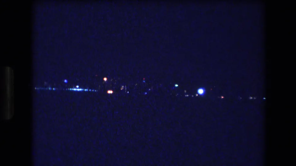 A handheld 8mm frame from Honolulu in 1977 shows brighter shoreline lights across dark water, with blue night exposure, visible grain, and scan banding.