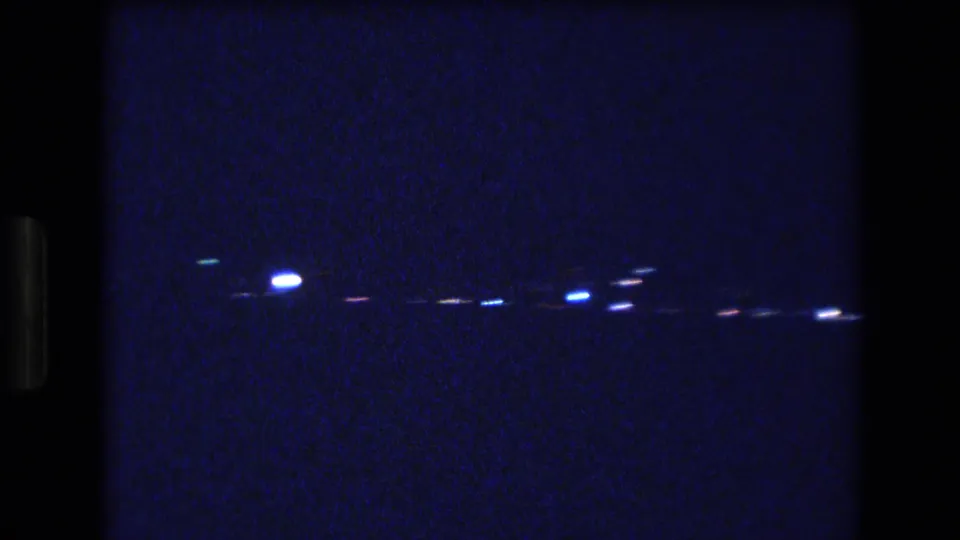 A late frame in the 1977 Honolulu night clip shows blurred blue-white lights along a dark horizon, softened by handheld motion and grainy 8mm exposure.