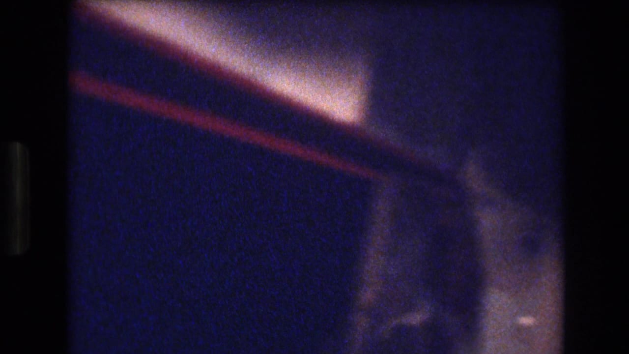 Soft, blue-tinted interior frame from the Honolulu party footage, showing a warm angled light or ceiling edge with indistinct room detail.