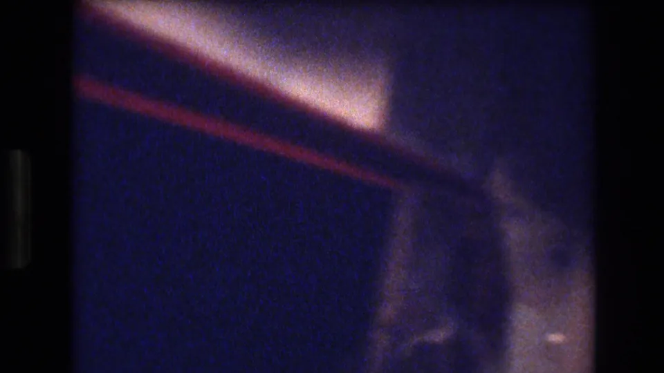 Soft, blue-tinted interior frame from the Honolulu party footage, showing a warm angled light or ceiling edge with indistinct room detail.