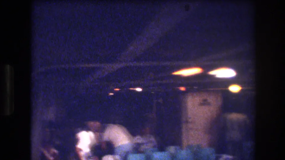 Grainy 8mm view of a dim 1977 Honolulu indoor party, with several people gathered near a doorway or corridor under warm ceiling lights.