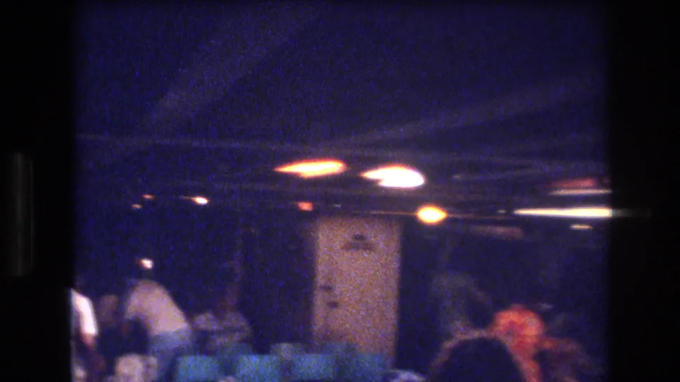 Final frame of the supplied 1977 Honolulu home-movie party clip, showing a crowded dim interior with guests near a doorway and warm overhead lighting.