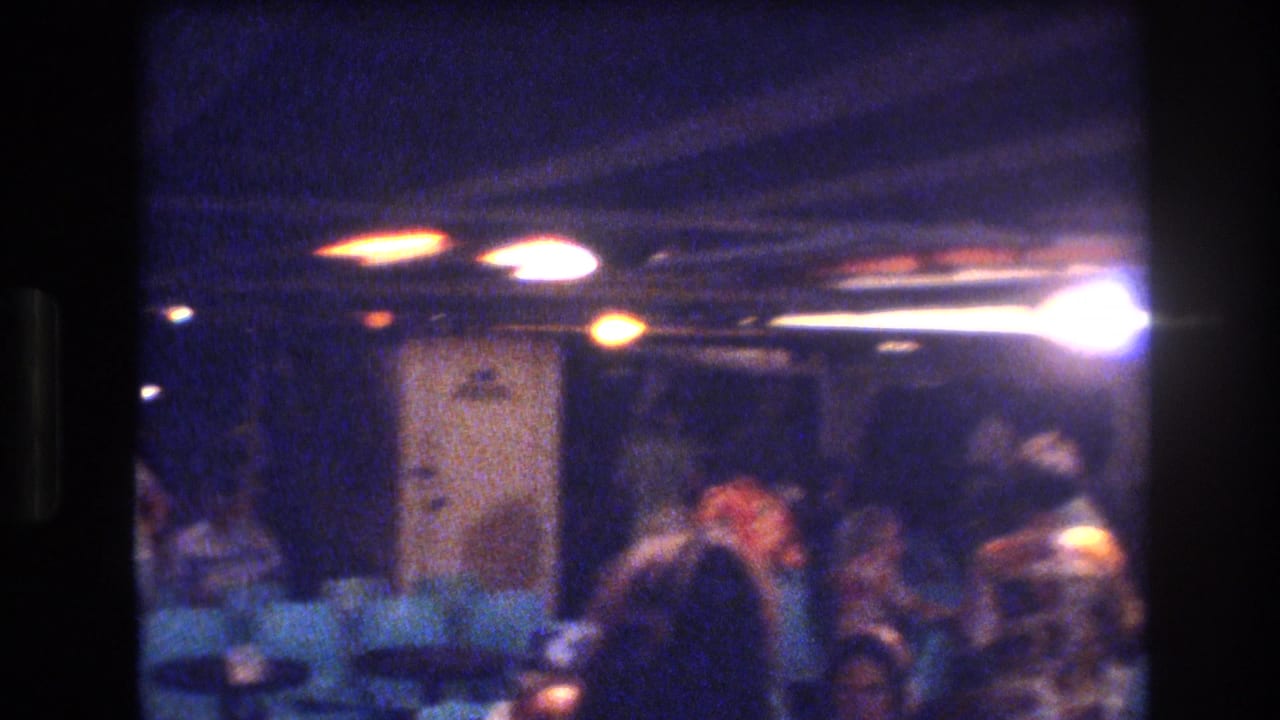 1977 Honolulu home-movie frame showing a dark blue-purple interior with adults gathered near teal seating and glowing overhead lights.