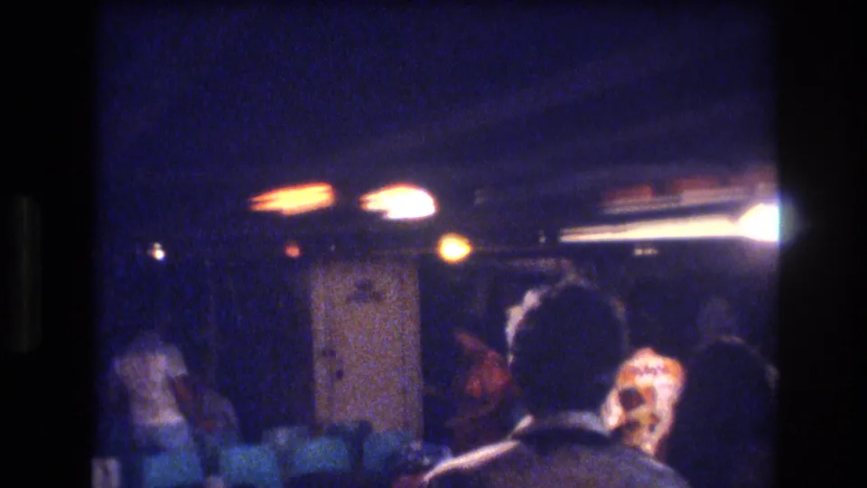 1977 Honolulu interior frame with adults moving or standing near seating in a low-lit room, filmed with soft focus and blue-purple color shift.