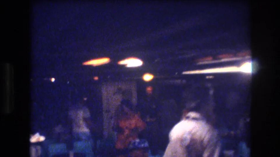 1977 Honolulu home-movie frame showing a dim social interior with blurred adult figures, teal chairs, and warm overhead light streaks.