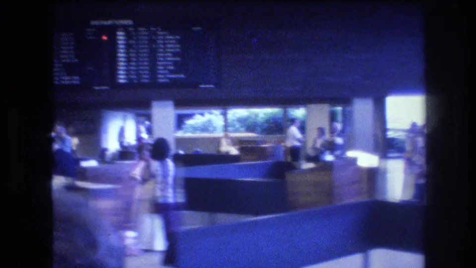 1977 Honolulu frame showing adults in a larger blue-lit interior with counters, seating, and an overhead information or display board.